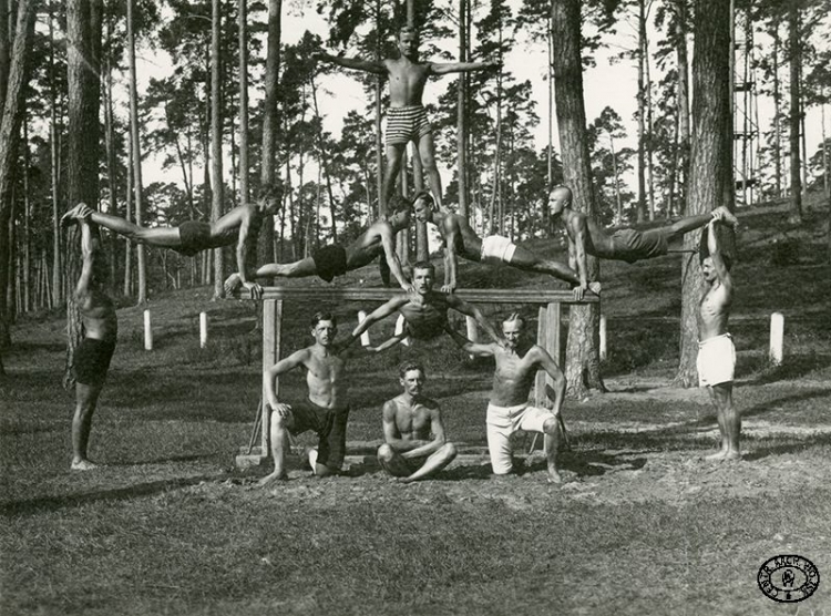 Oficerowie legionowi podczas ćwiczeń akrobatycznych w obozie internowanych. Od lewej pod poprzeczką: por. Stanisław Łapiński „Nilski”, por. Jan Lewandowski (siedzi), ppor. Ludwik Skrzyński „Kmicic” (na górze), kpt. Stefan Biernacki „Dąb”; bokiem od lewej: kpt. Władysław Orzechowski „Dragat”, ppor. Jan Sadowski „Jagmin” (w spodenkach w pasy), rtm. Wacław Graba-Łęcki (piąty). Beniaminów. Lato 1917 r.? Źródło: CAW