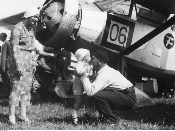 Porucznik pilot Franciszek Żwirko wita się ze swoim synkiem Heniem i żoną Agnieszką po przylocie na warszawskie lotnisko w sierpniu 1932 r.. Z tyłu widoczny samolot RWD-6. Fot. Narodowe Archiwum Cyfrowe.